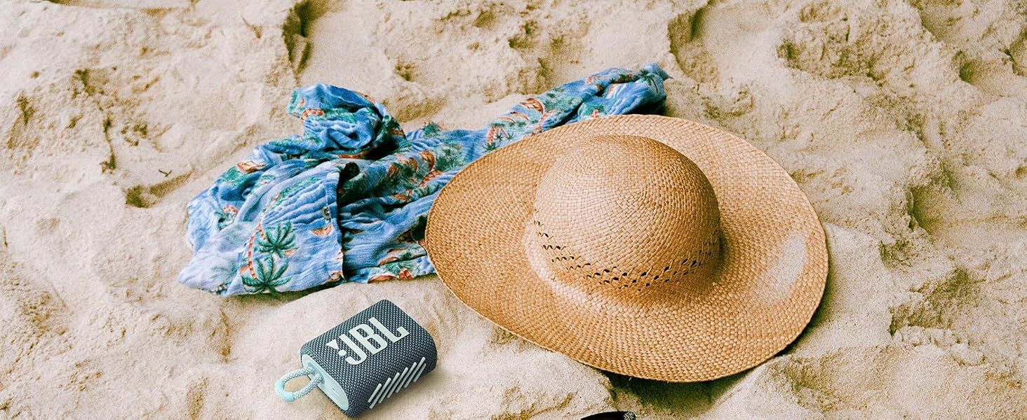 Shows the JBL speaker resting on sand beside a straw hat and scarf to help shoppers envision using it on a beach day outdoors.