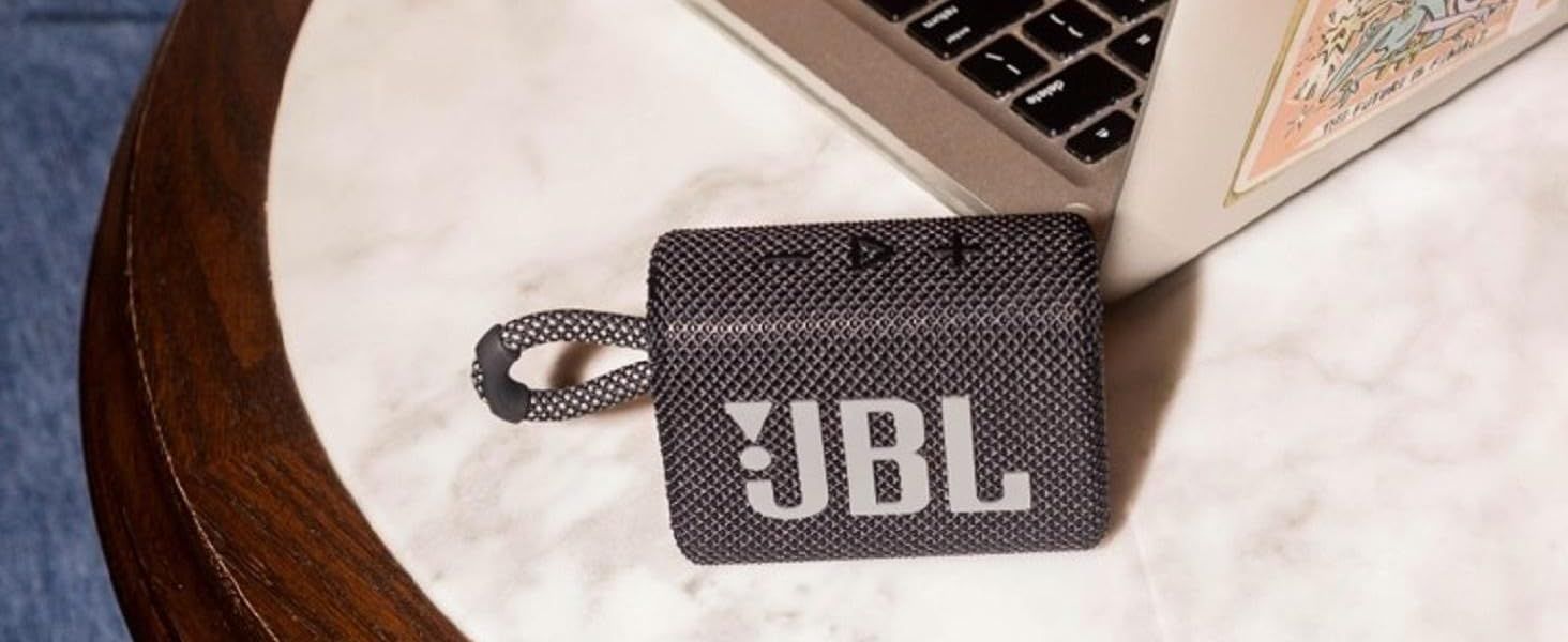 Places the JBL logo front and center on a lifestyle scene so shoppers recognize the brand and associate the speaker with its established identity. Shows the compact speaker resting on a marble table…