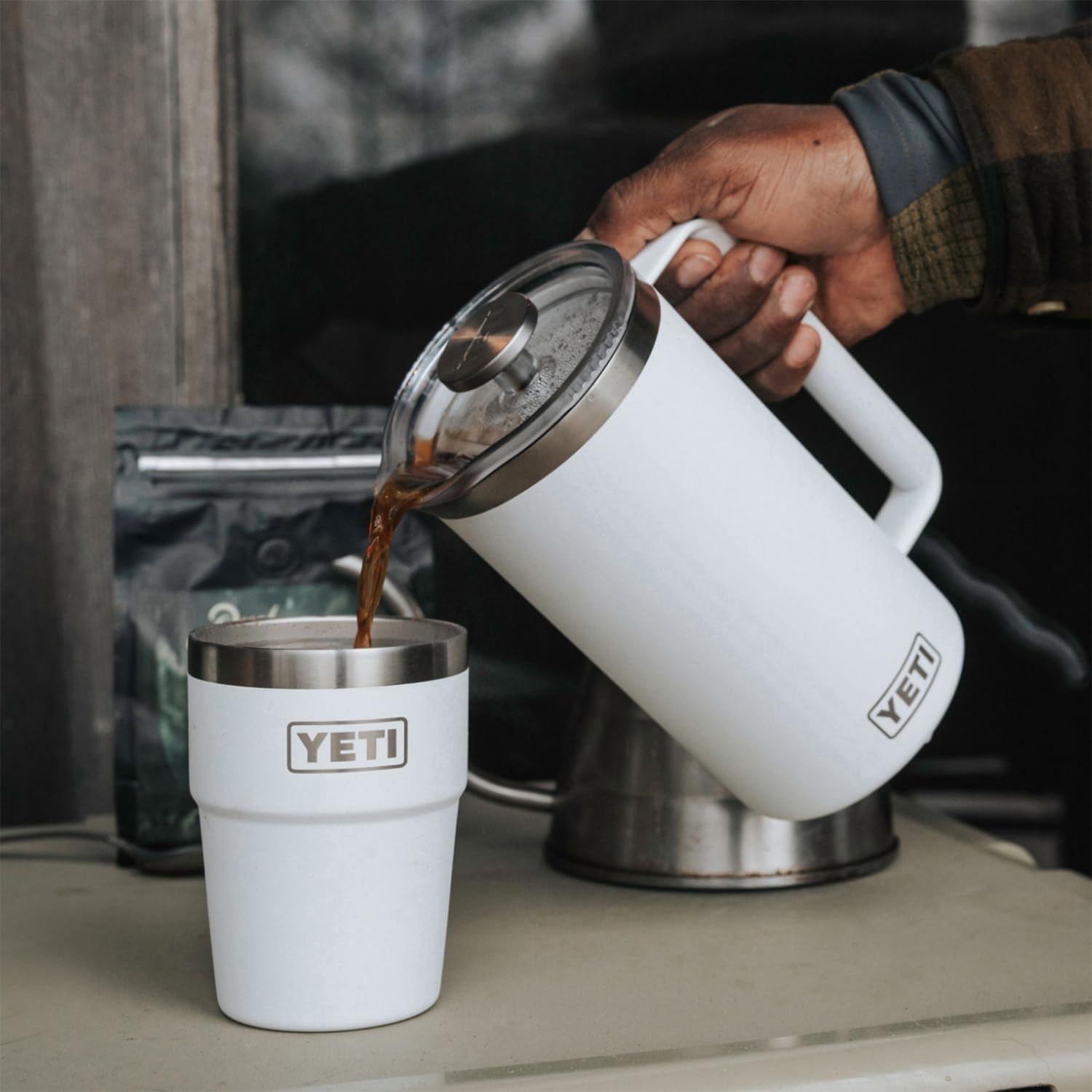 Demonstrates the product in use by showing someone pouring coffee from the press into a tumbler in a real-world setting.