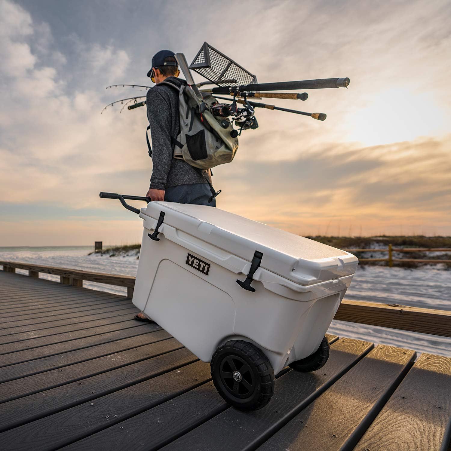 Shows the cooler in a real-world fishing context on a dock with a person carrying fishing gear, demonstrating the product's use in outdoor recreational settings without any text overlay.