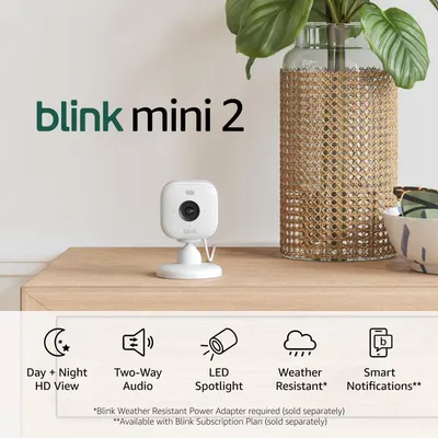 Shows the Blink Mini 2 camera in a real home environment to help shoppers visualize how it fits naturally into their living space. Uses graphic overlays including the product name and feature icons…