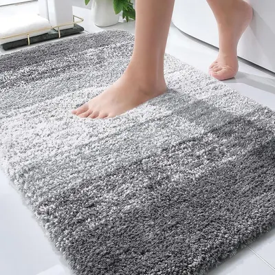 Shows a person standing on the bath mat to demonstrate its size, texture, and real-world use, helping shoppers visualize how it fits in their bathroom space. Displays the bath mat in a realistic…