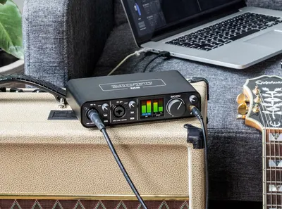 Shows the audio interface in an authentic home studio environment with guitar amp and laptop to help shoppers visualize how the product fits into their creative workflow.