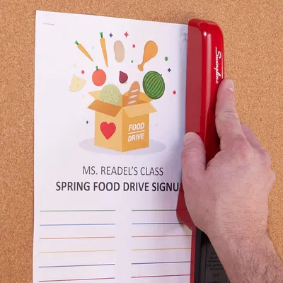 This is not an Amazon product image but rather a photo of a stapler being used on a food drive signup sheet. The image shows the product (red Swingline stapler) in a real-world context on a cork…