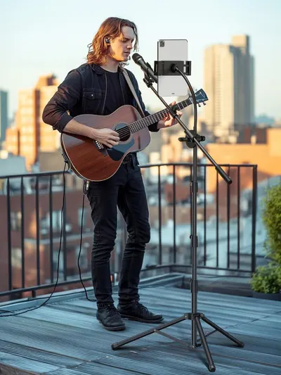 Demonstrates the product in a real-world use case, showing a musician using the microphone and tablet stand setup for outdoor recording or live streaming on a rooftop, emphasizing versatility and…