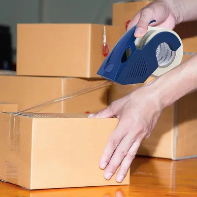 Demonstrates the product in a real-world context, showing someone using the tape dispenser to seal cardboard boxes, helping shoppers visualize practical everyday use.