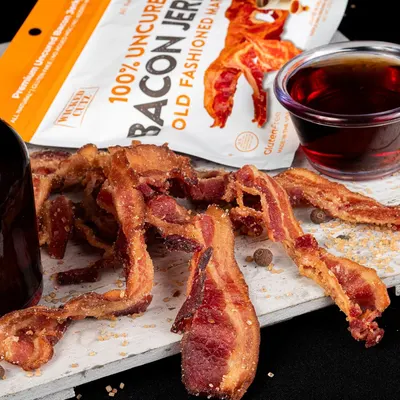 Showcases the bacon jerky product in an appetizing, real-world setting with complementary beverage to help shoppers visualize consumption and appeal.