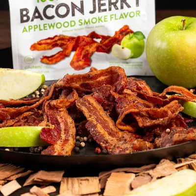 Showcases the bacon jerky product in an appetizing, real-world serving context with fresh apples and peppercorns to help shoppers visualize how they might enjoy the product.