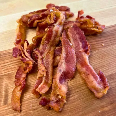 Shows the product (bacon) in a real-world context on a wooden cutting board to help shoppers visualize the cooked result and quality of the product.