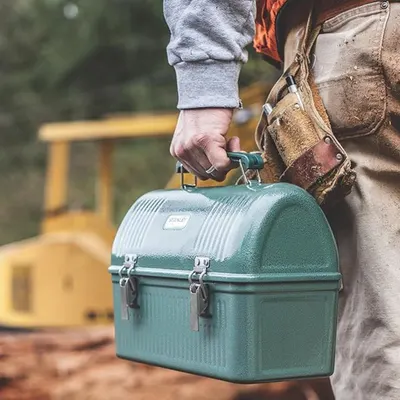 Depicts the lunch box being carried by a worker in a construction or work environment, highlighting its durability and practical use for tradespeople.