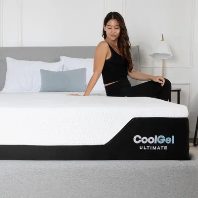 Displays the mattress in a real bedroom environment with a person sitting on it, helping shoppers visualize the product in their own home.