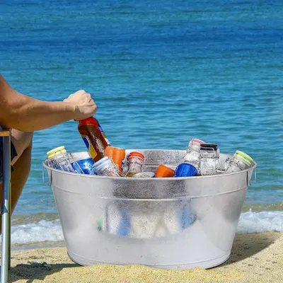 Shows the galvanized beverage tub filled with drinks on a sunny beach, conveying a relaxed outdoor lifestyle without any text overlay.